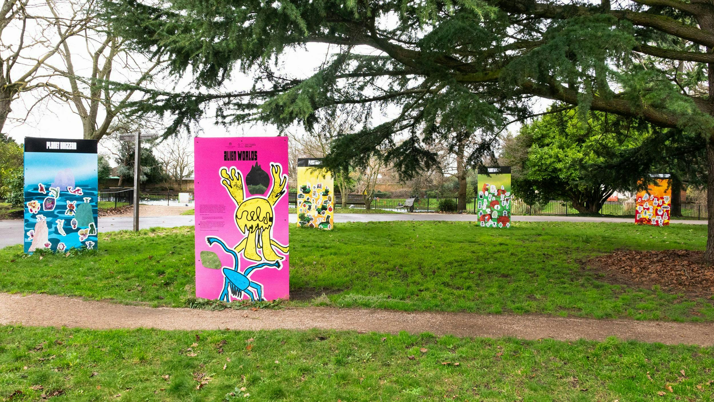Five large colourful signs with drawing on, in a park with large tree branch in foreground